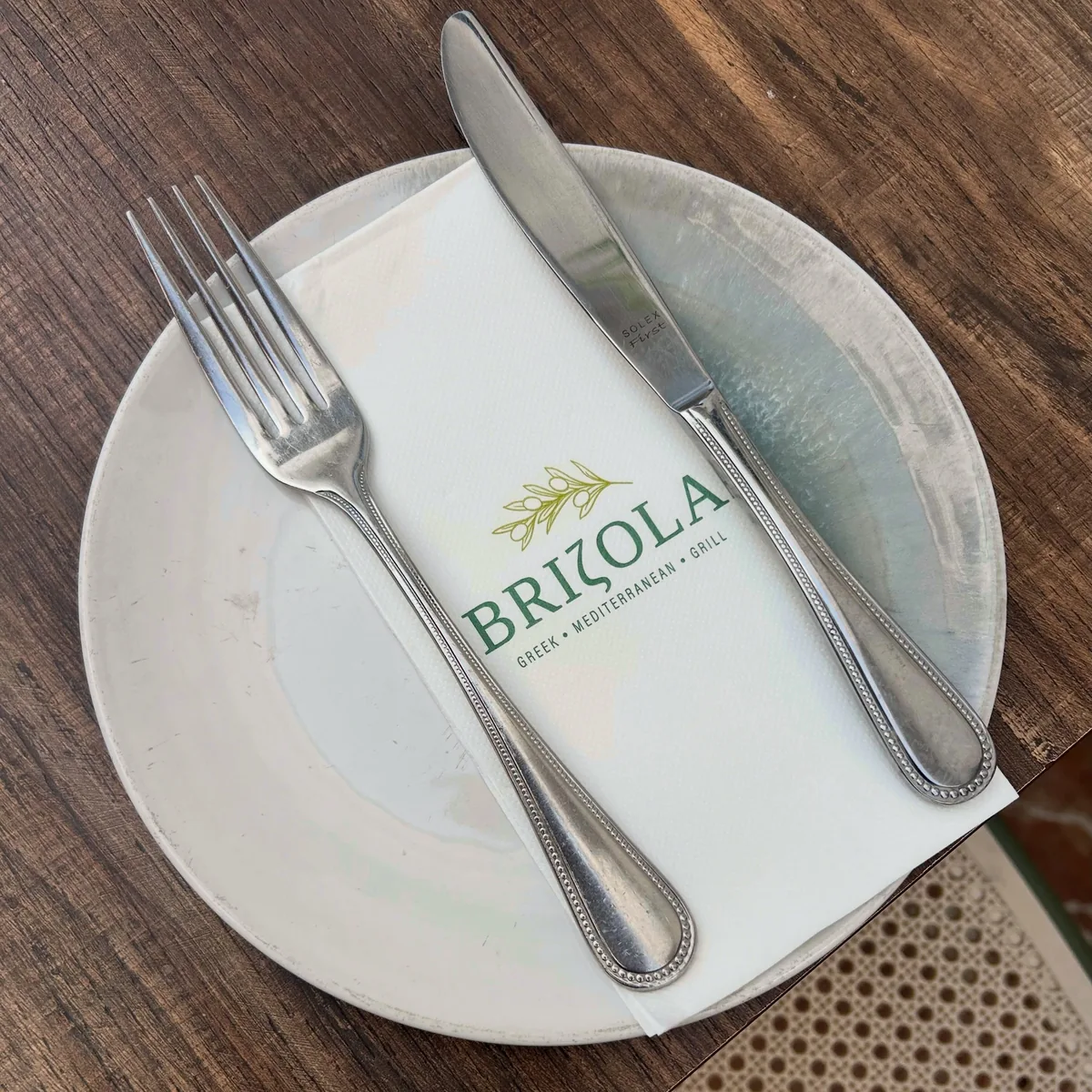 A Brizola place setting — white ceramic plate, silver cutlery and a napkin printed with the olive-branch wordmark