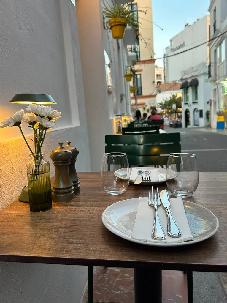 A laid table on the Brizola terrace at dusk, with an olive-green lamp, daisies and the Estepona street beyond